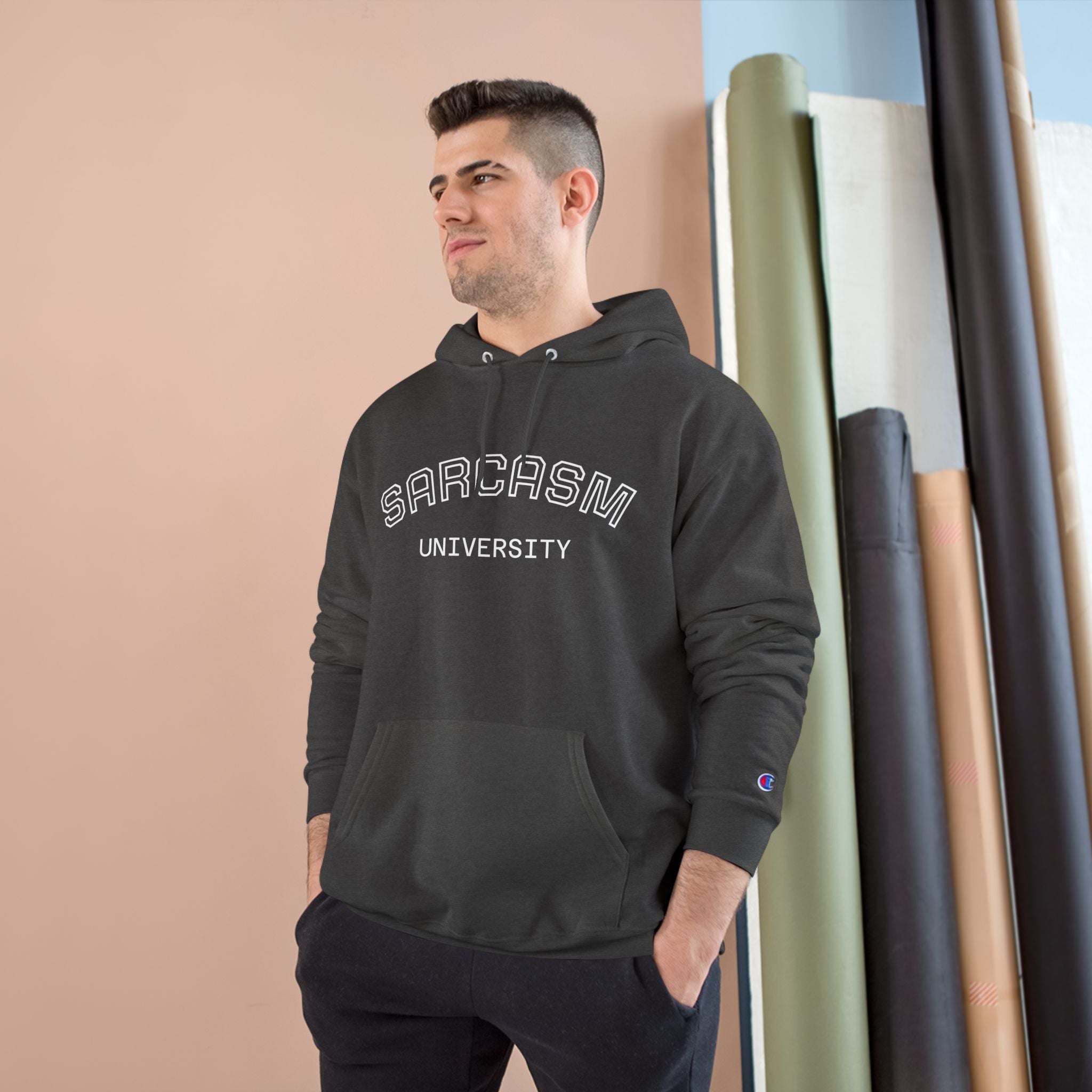 Man wearing a black hoodie with 'SARCASM UNIVERSITY' text, standing against a beige wall.