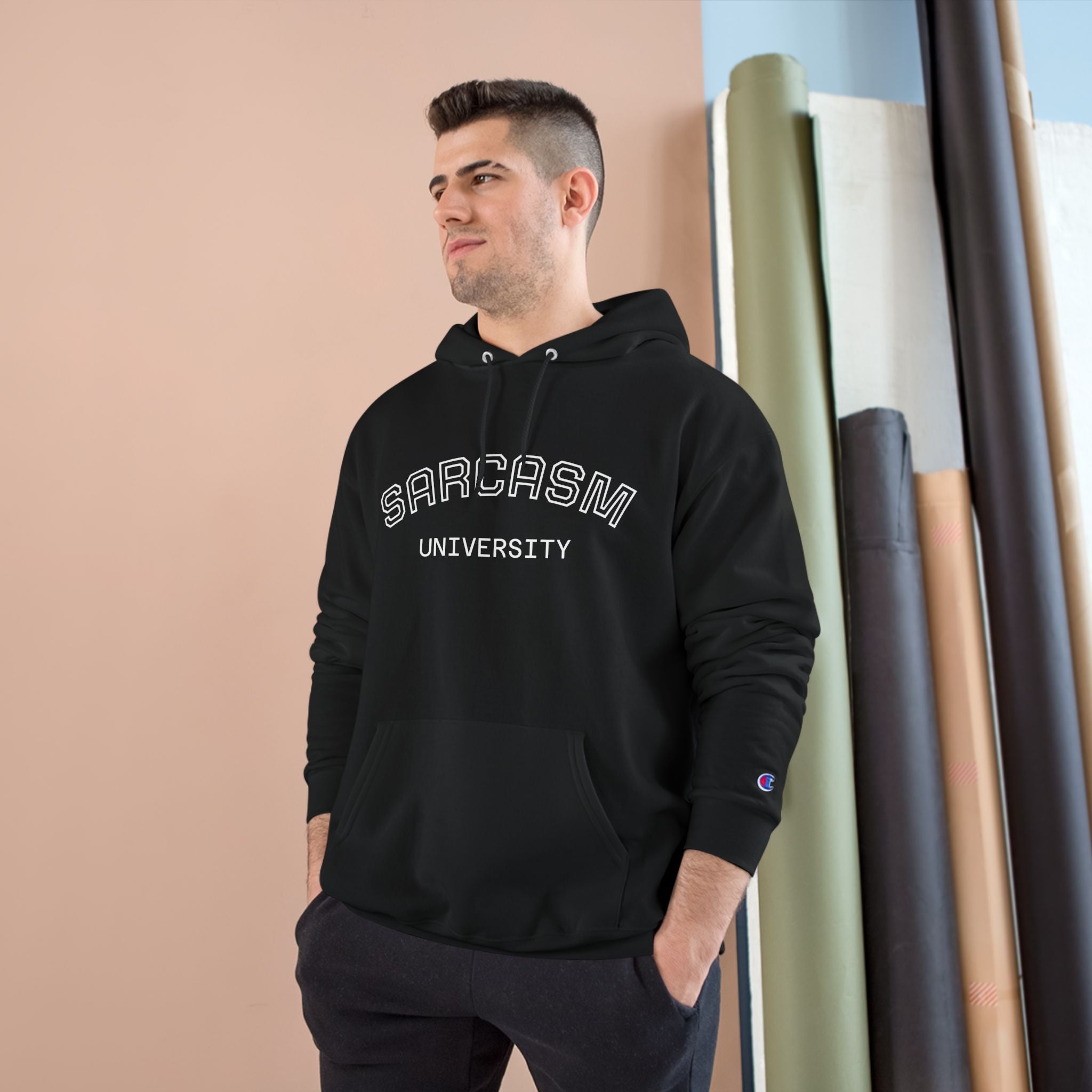 Man wearing a black hoodie with 'SARCASM UNIVERSITY' text, standing against a beige wall with art supplies.