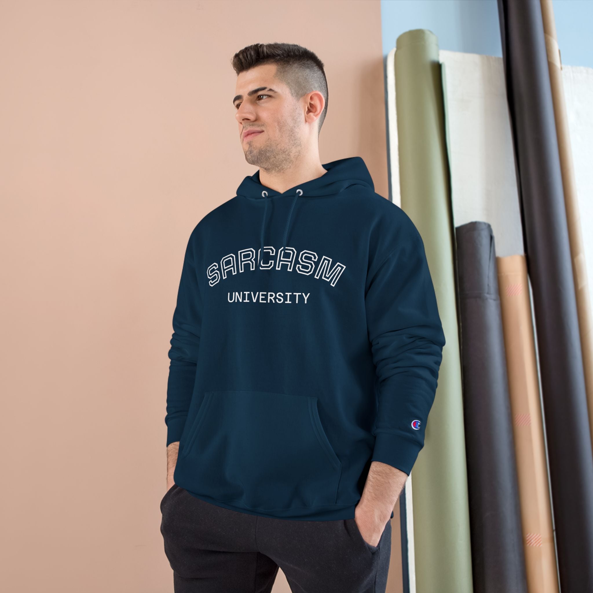 Man wearing a navy hoodie with 'SARCASM UNIVERSITY' text, standing against a beige wall with art supplies.